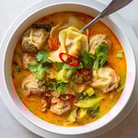 A bowl of steaming Red Curry Wonton Soup With Greens, garnished with fresh cilantro and lime wedges.